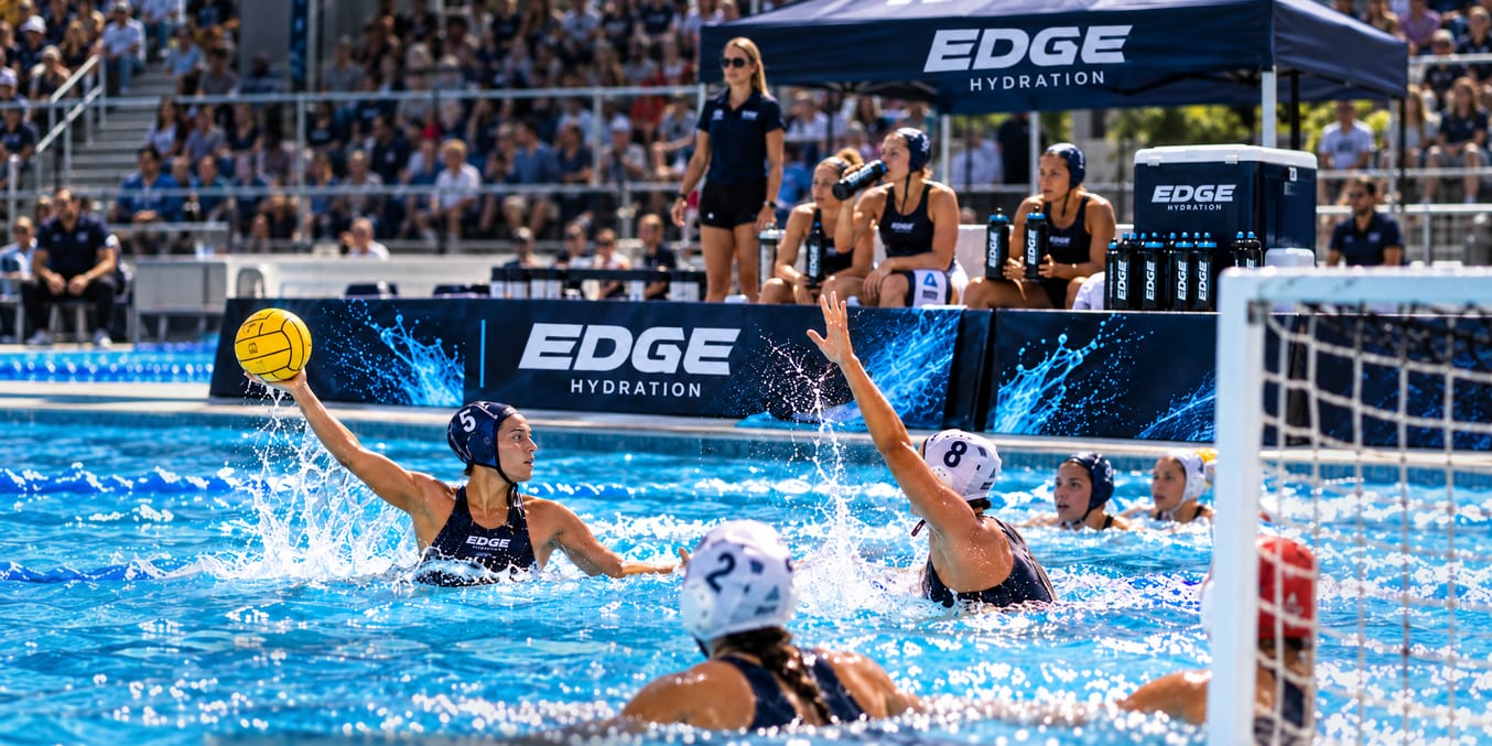 Women’s water polo match beside EDGE Hydration branding, with players on the bench and fans in the stands.